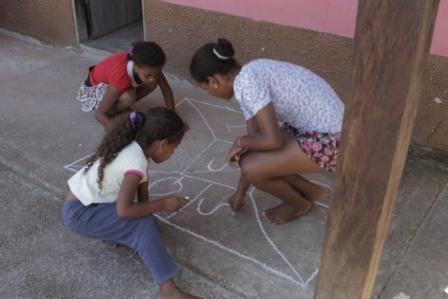 Foto com três meninas ambas estão abaixadas, em circulo elas desenham em uma calçada com giz de lousa.