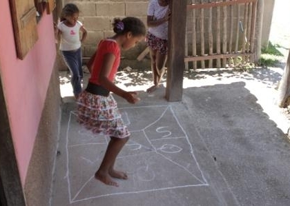 Foto com três crianças brincando, uma esta pulando amarelinha enquanto as outras duas observam.
