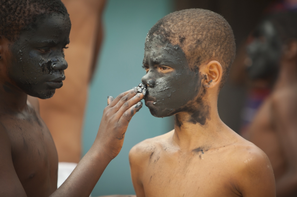 Foto composta por dois garotos, onde o primeiro garoto negro está com a cara cheia de lama, passa lama na cara do segundo garoto com uma expressão séria.