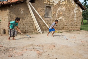 Foto com dois garotos brincando, com um eixo com duas rodas presas em suas extremidades, ele empurra com um graveto que está preso ao eixo.