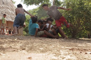 Foto de crianças sentadas em fila indiana, duas crianças correm em volta delas.