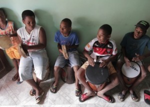 Foto onde cinco crianças estão tocando instrumentos, três estão tocando pandeiro e duas estão tocando tambor.