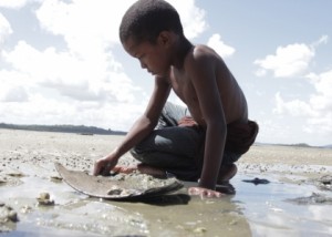 Foto com um garoto negro sem camisa ajoelhado, mexe em um chão lamacento.