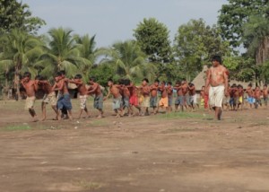 Foto com uma fila com crianças andam em fila indiana, algumas com as mão no ombro do próximo e outra com as mãos na cintura, um homem anda ao lado das crianças.