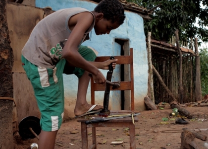 foto de um garoto negro que esta encurvado com seu pé esquerdo sobre a cadeira, com seu braço esquerdo apoia um facão em um pedaço de madeira em cima da cadeira, e com seu braço direito segura um martelo.