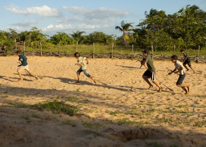 Foto composta por crianças correndo em um chão de areia, ambas as crianças estão com um galho de folha de um coqueiro entre as pernas, como se fossem cavalos.