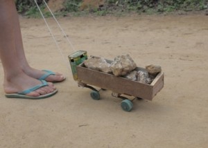 Foto ilustrando um caminhão de brinquedo feito de madeira carregando pedras, um garoto segura uma corda que está amarrado a este caminhão.