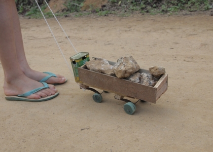 Foto ilustrando um caminhão de brinquedo feito de madeira carregando pedras, um garoto segura uma corda que está amarrado a este caminhão.