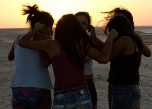Foto ilustrando cinco garotas em círculo ao pôr do sol, cada uma delas estão com as mãos nas orelhas das pessoas ao seu lado uma mão, em cada pessoa.