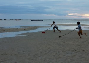 Foto demonstra dois garotos correndo na praia ao entardecer, cada um empurra seu brinquedo com um graveto que está amarrado a ele, o brinquedo é um eixo de madeira com carretéis em suas extremidades.