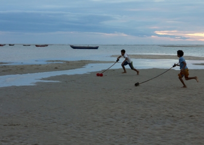 Foto demonstra dois garotos correndo na praia ao entardecer, cada um empurra seu brinquedo com um graveto que está amarrado a ele, o brinquedo é um eixo de madeira com carretéis em suas extremidades.