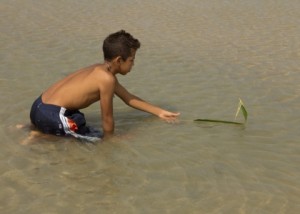 Foto de um garoto ajoelhado brincando dentro de um lago mexe em uma folha que esta boiando.