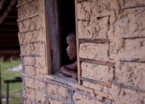 Foto com um garoto negro encostado em uma janela dentro da sua casa, observa fora.