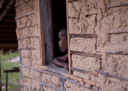 Foto com um garoto negro encostado em uma janela dentro da sua casa, observa fora.