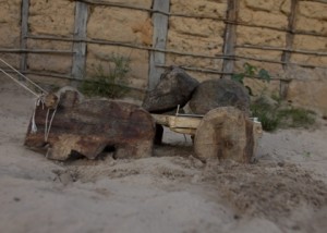 Foto de um brinquedo de um boi que puxa uma carroça feito de madeira, tem duas pedras em cima da carroça do boi, o brinquedo está sendo puxado com um barbante.