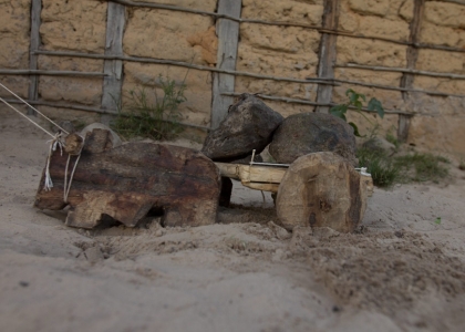 Foto de um brinquedo de um boi que puxa uma carroça feito de madeira, tem duas pedras em cima da carroça do boi, o brinquedo está sendo puxado com um barbante.