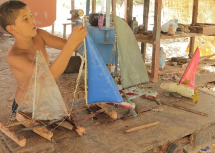 Foto de um garoto mexendo em um barco a vela de brinquedo que está em cima da mesa, tem quatro barcos em cima da mesa.