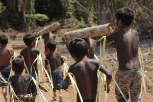 Foto composta por varias crianças andando com pinturas em seu corpo, algumas delas carregam troncos de madeira.