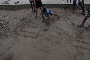 Foto de crianças brincando com uma tampinha em um chão de terra.