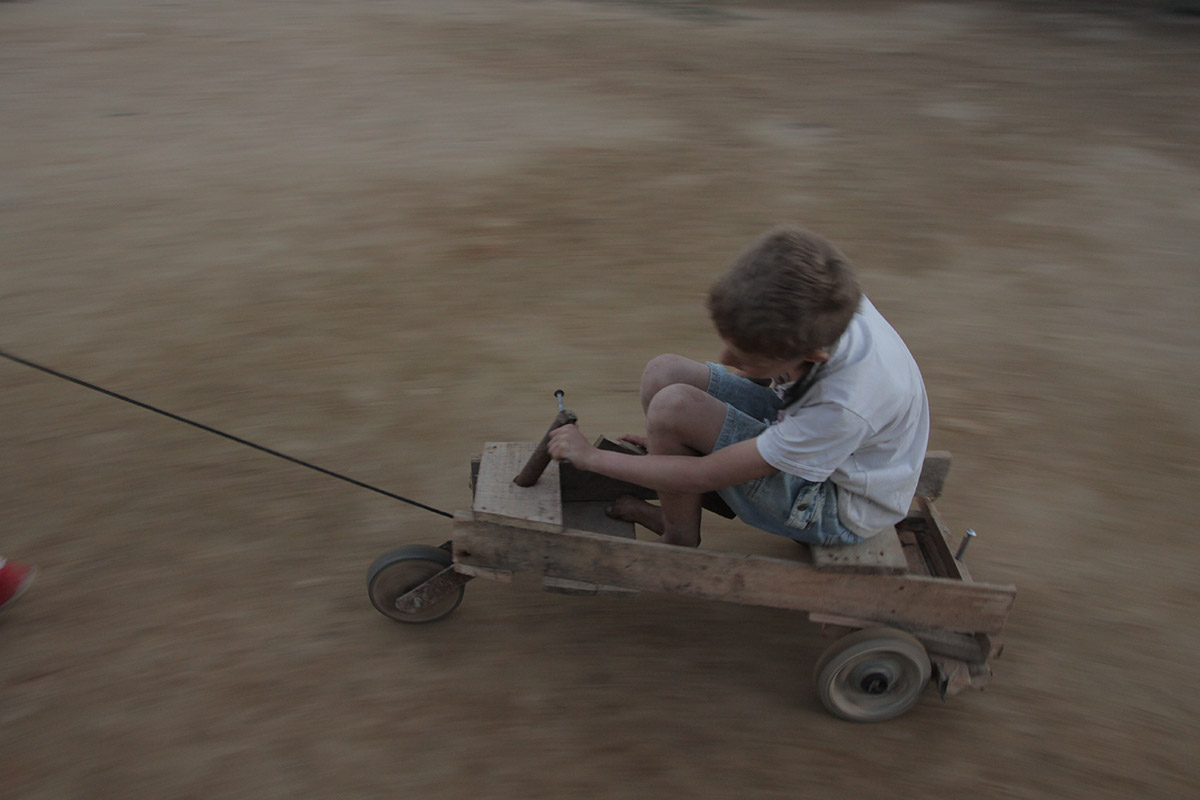 Foto com um garoto brincando em cima de um carrinho de madeira está sendo puxado por um cabo amarrado ao carrinho.