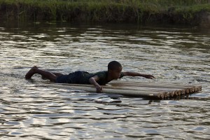 Foto de um garoto deitado de barriga em uma placa feita com galhos de galhos de folhas de bananeira, o garoto está remando com seus braços.