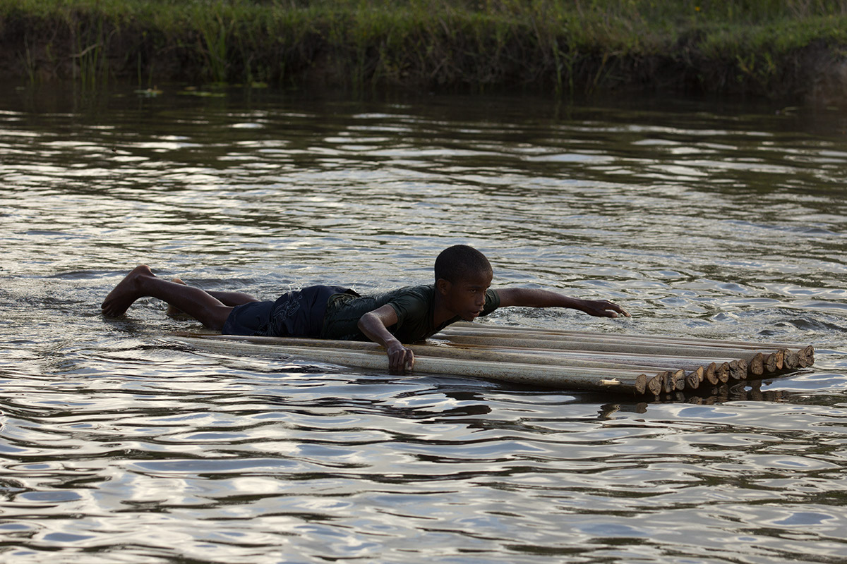 Foto de um garoto deitado de barriga em uma placa feita com galhos de galhos de folhas de bananeira, o garoto está remando com seus braços.