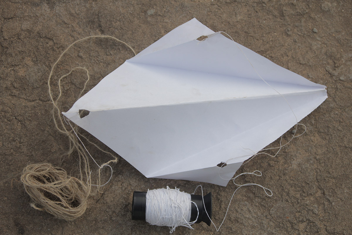 Foto de um pipa feita de folha sulfite em um chão de terra, que amarrado a ele tem um carretel de barbante.