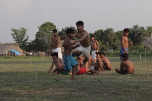 Foto com várias crianças sentadas e quatro homens em pé, dentro de uma cerca de barbante bem baixa ao chão, um dos homens estica seus braços para pegar um garoto que está correndo em volta da cerca.