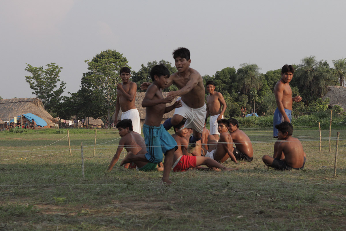 Foto com várias crianças sentadas e quatro homens em pé, dentro de uma cerca de barbante bem baixa ao chão, um dos homens estica seus braços para pegar um garoto que está correndo em volta da cerca.
