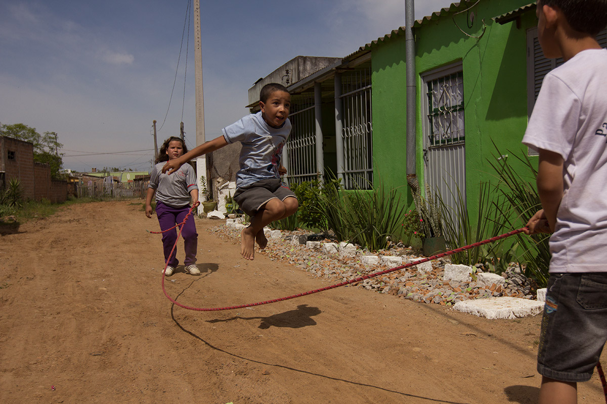 Foto demonstra três crianças brincando de pular corda, um garoto esta saltando, enquanto outras duas crianças, um menino e uma menina, estão batendo a corda.