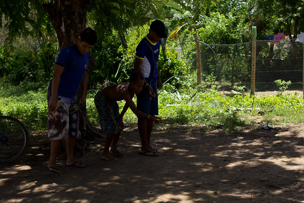 Foto com três garotos brincando de bolinha de gude em baixo de uma arvore.