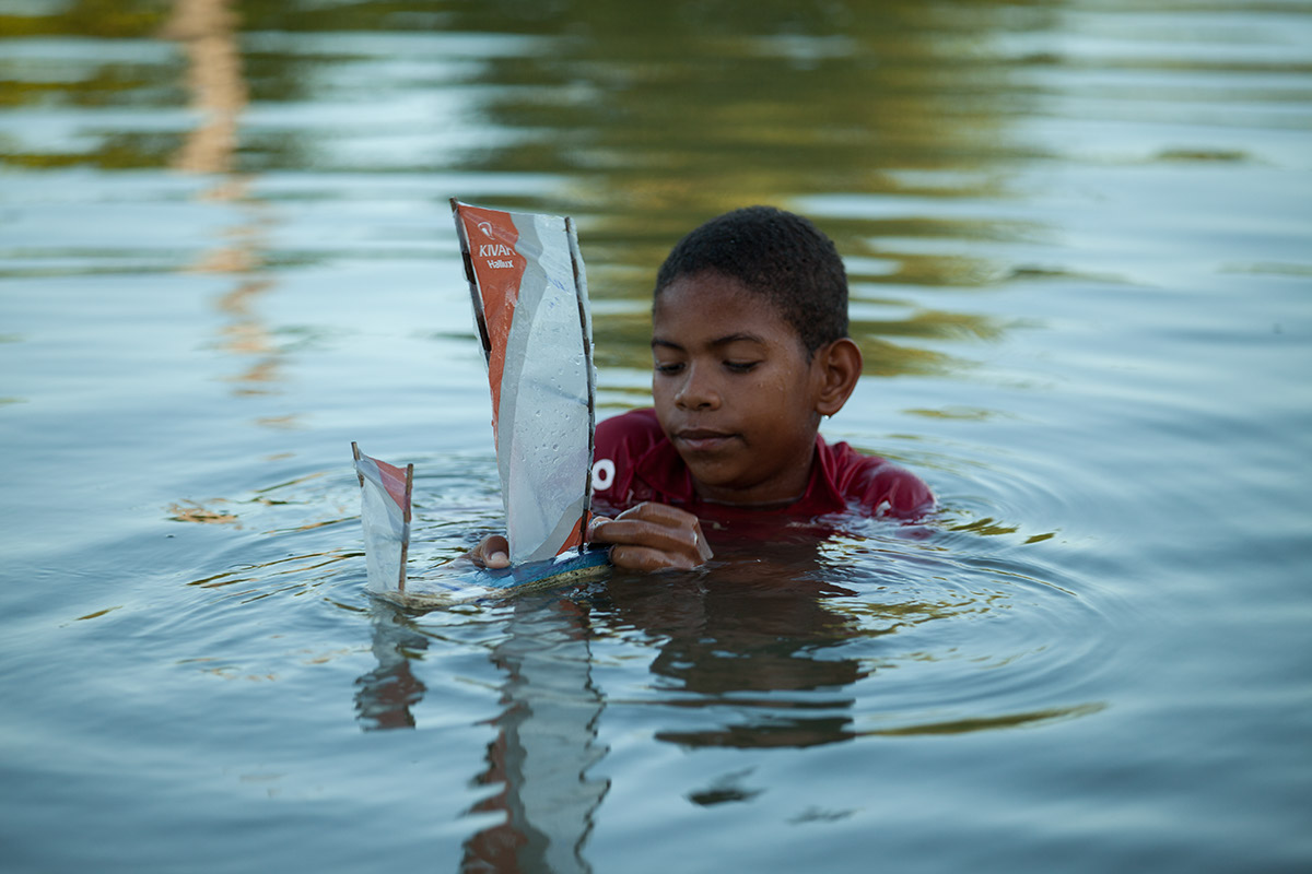 Foto de um garoto dentro de um lago, brinca com um barco a vela de brinquedo.
