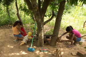 Foto com duas meninas brincando de casinha ambas abaixadas uma mexe em um saco vermelho enquanto outra mexe em uma mesa de brinquedo feito de madeira com alguns utensílios, vários brinquedos estão espalhados.