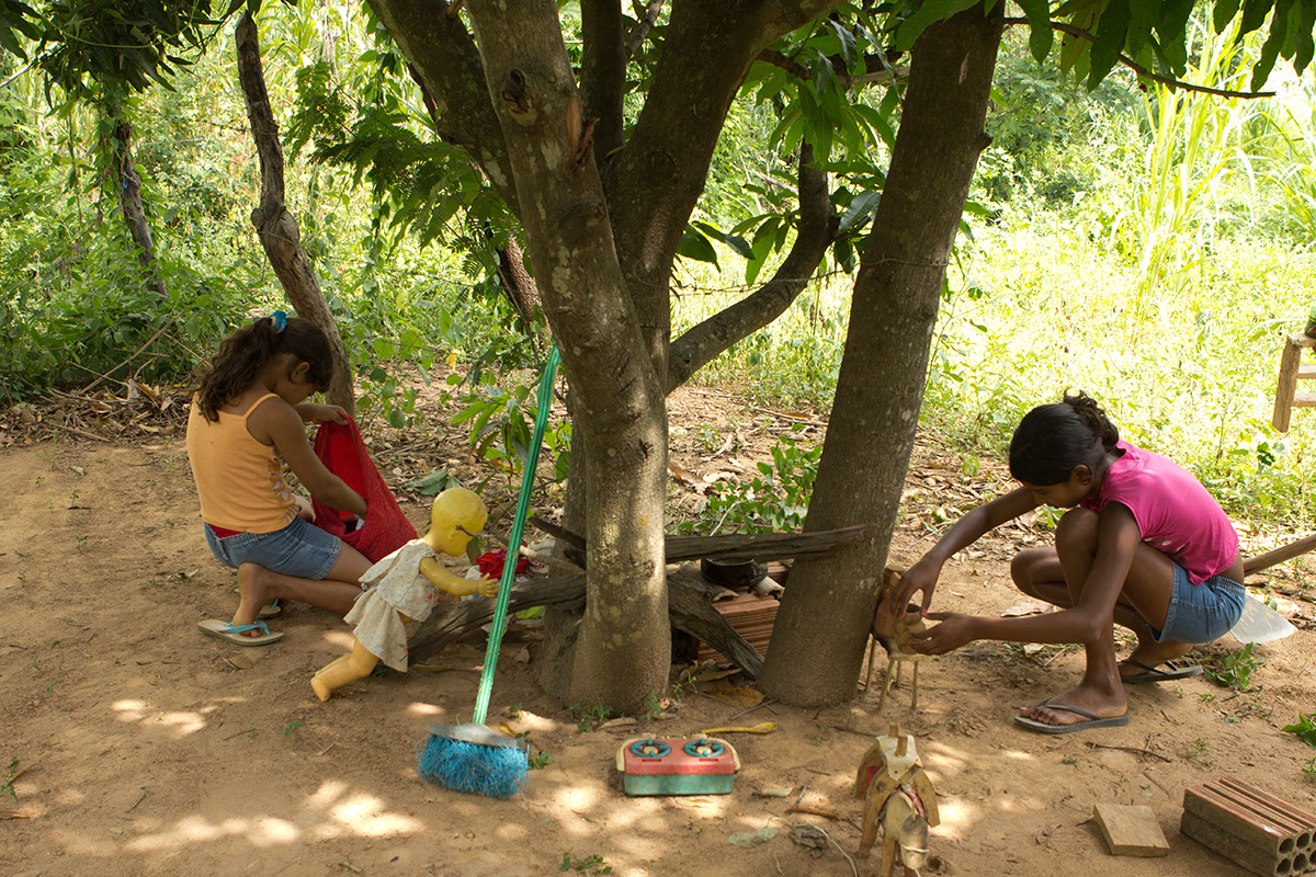 Foto com duas meninas brincando de casinha ambas abaixadas uma mexe em um saco vermelho enquanto outra mexe em uma mesa de brinquedo feito de madeira com alguns utensílios, vários brinquedos estão espalhados.