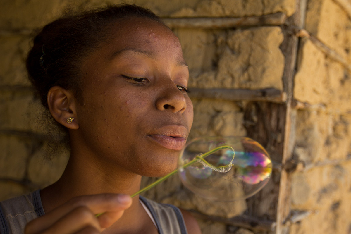 Foto de uma garota que segura uma folha que em sua ponta esta dobrada, formando um aro, ela utiliza deste aro para assoprar e fazer bolinhas de sabão.