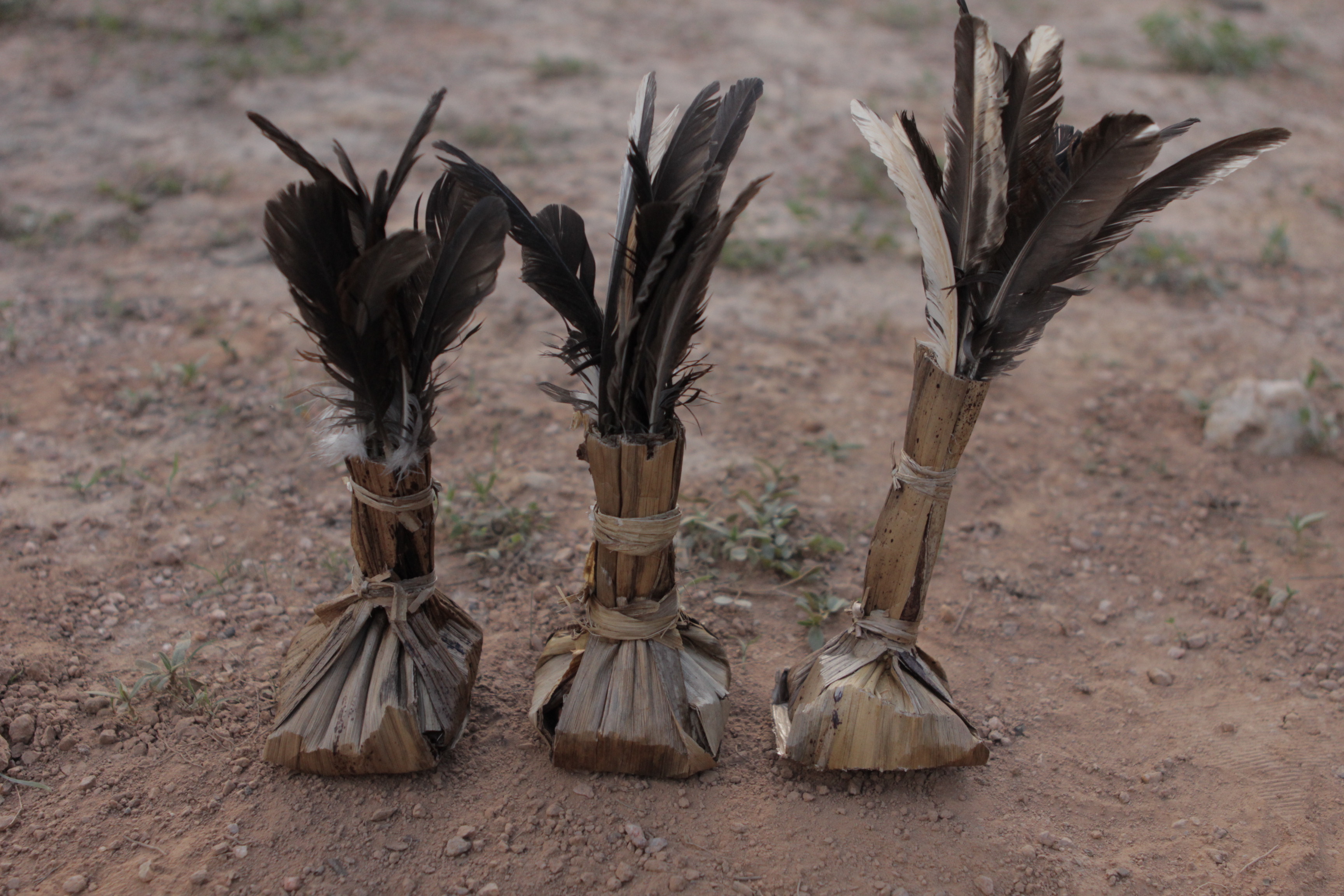 Foto de tres petecas feitas com palha e pena em um solo de terra.