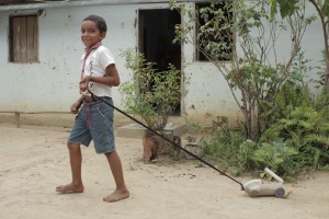Foto com um garoto puxando um carrinho feito de uma garrafa de alvejante, o garoto puxa com uma corda amarrada.