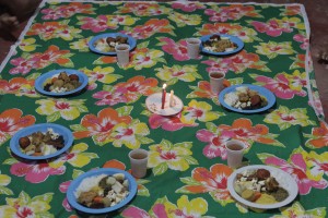 Foto de um pano multicolorido estendido, que em cima dele tem vários pratos plástico com comida dentro e um copo de plástico com suco ao lado deles, os pratos formam um círculo, no centro tem um prato com três velas acessas.