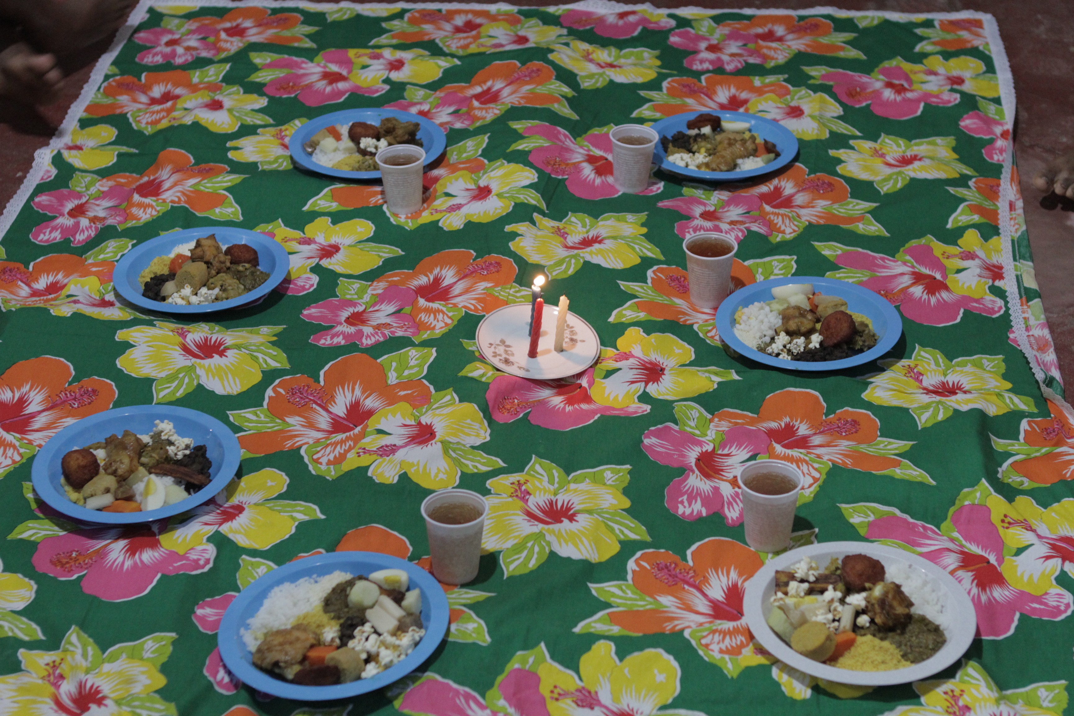 Foto de um pano multicolorido estendido, que em cima dele tem vários pratos plástico com comida dentro e um copo de plástico com suco ao lado deles, os pratos formam um círculo, no centro tem um prato com três velas acessas.