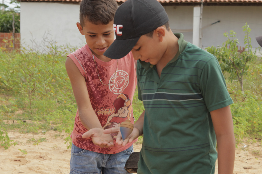 Foto com duas crianças brincando um peão, as duas crianças estão com a mão direita estendida, um garoto de blusa vermelha e outro de boné preto, o garoto de boné preso segura um peão que gira em sua mão.