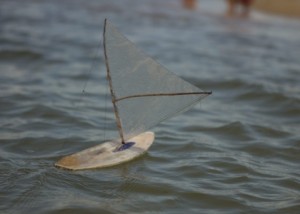 Foto de um barquinho a vela feito com plástico linhas e madeira em um rio.