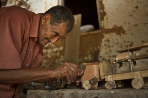 Foto de um homem negro idoso, mexe em um caminhão de brinquedo feito de madeira carregando carrinhos em sua carga.