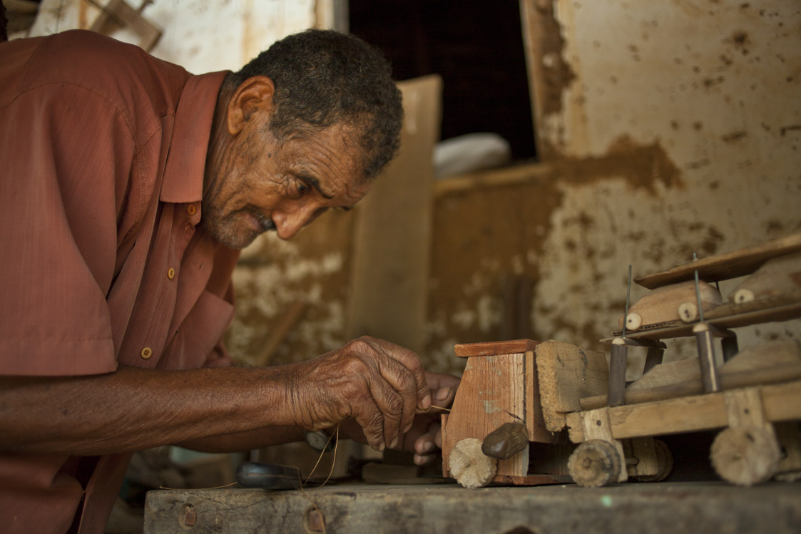 Foto de um homem negro idoso, mexe em um caminhão de brinquedo feito de madeira carregando carrinhos em sua carga.