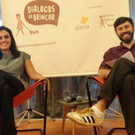 Foto com duas pessoas sentadas, uma mulher e ao seu lado esquerdo um homem de barba, ambos estão de pernas cruzadas e sorrindo.