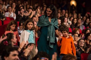 Foto de um salão cheio de pessoas sentadas observando uma mulher descer as escadas, de mãos dadas a uma menina e um menino.