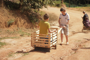 Foto de dois garotos brincando com um carrinho de madeira , onde um dos garotos está dentro do carrinho e o outro está do lado de fora puxando o carrinho de madeira.