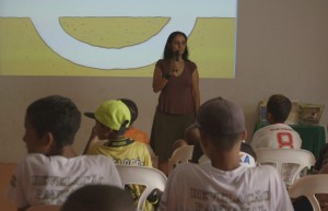 Foto de um salão composta por várias pessoas, uma mulher em frente a um telão, fala ao microfone para uma turma que está sentada e a observa.