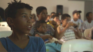 Foto composta por várias crianças sentadas, elas estão a observar algo.