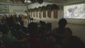 Foto compostas por varias pessoas em um salão, olhando para um telão que esta sendo projetado um filme.