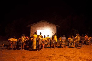 Foto de um grupo de pessoas de costas para a foto, observam ao fundo um telão sendo projetado em uma parede de uma casa de tijolos.
